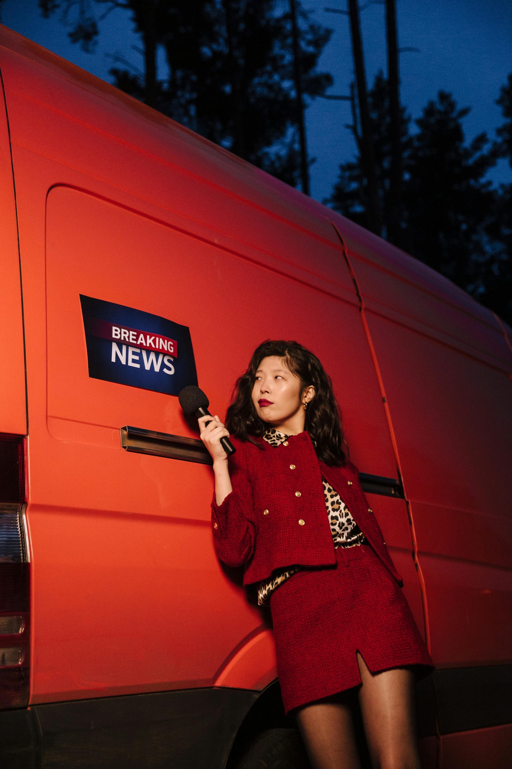 Asian woman reporter in red standing by a breaking news van at twilight.